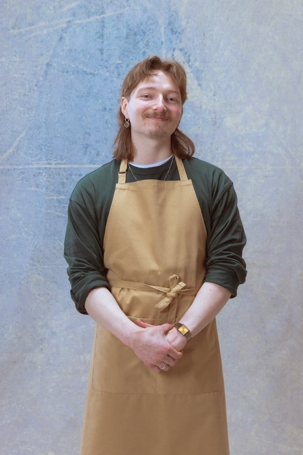 A man wearing a beige apron, standing with his hands clasped and smiling at the camera