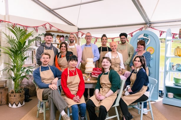 12 people sitting in the Great British Bake Off Tent smiling at the camera