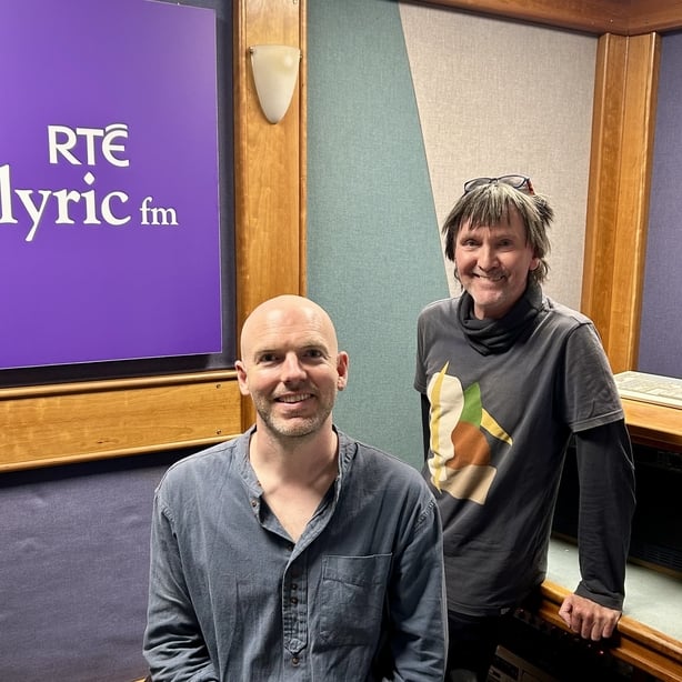 Michael Gallen seated in front of an RTE Lyric FM sign with Paul Herriott standing behind him