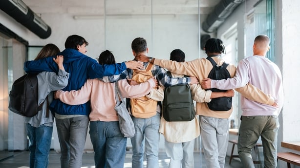 A group of diverse young students stands together, embracing each other warmly, symbolizing friendship, support, and camaraderie. They are in a bright, modern indoor setting, fostering unity and connection.
