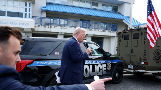 A man points as Donald Trump speaks into a microphone beside a police car 