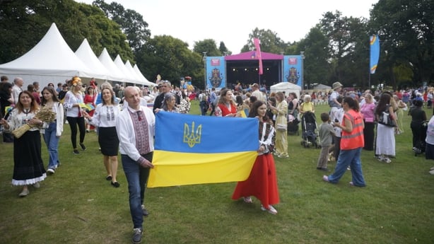 Crowds of people are seen at a festival with tents lined along the left hands side, and a blue and yellow flag in the centre