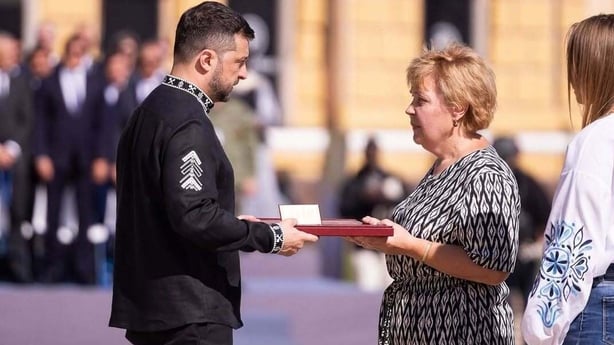 A man wearing black clothes and military emblem hands a medal to a woman wearing a black and white dress during a memorial service