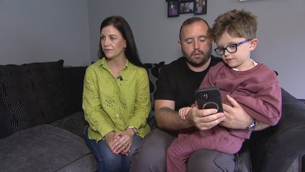 A man has his child on his knee as they both look down at a phone while a woman sits beside them staring off camera