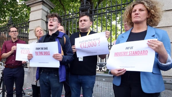 A group of people stand outside a a large gate holding signs that read 'could you live on €12,000'