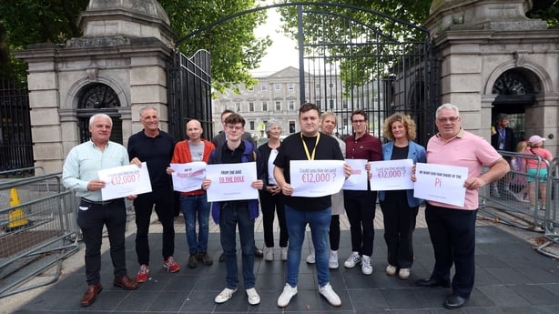 A group of people stand outside a a large gate holding signs that read 'could you live on €12,000'