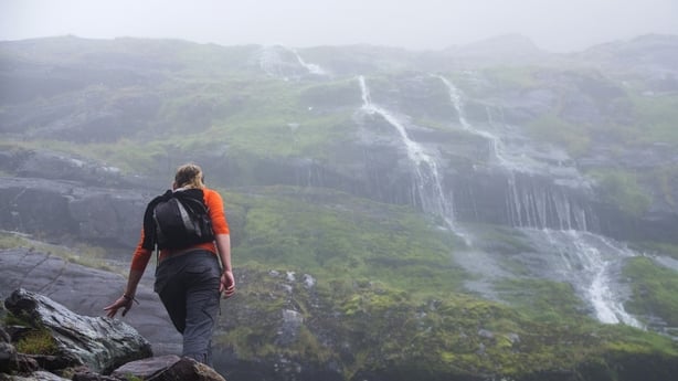 A woman with a small backpack and Orange stops for a rest while the water is pouring down the mountain nearby on Carrauntoohil in Killarney, Kerry in Ireland