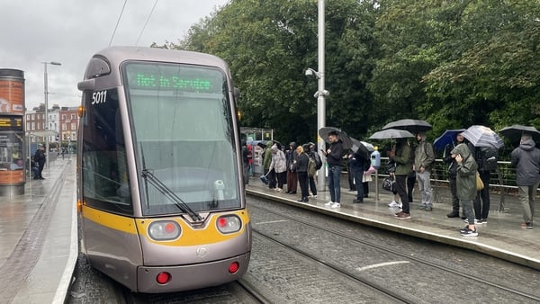 A luas tram at St Stephen's Green stop with a 'not in service' sign as people wait in the rain, some with umbrellas