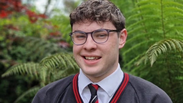 A young man in a school uniform poses for a photo in front of greenery.