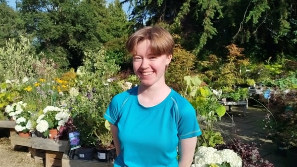 A young person in a blue t-short smiles as they pose in a garden.