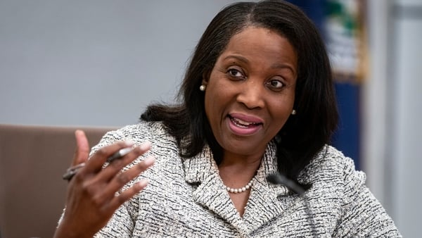 A woman in a light grey specked suit raises her hand at a meeting