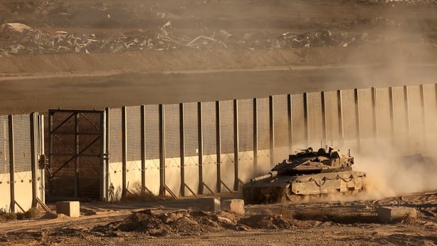 A tank moves along a border area next to a wall with dust trailing behind it