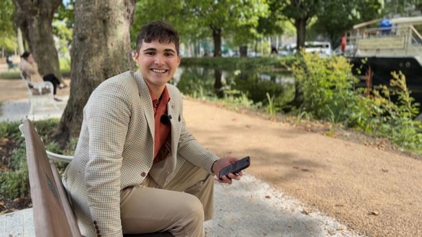 A young, smiling man wearing a cream jacket sits on a benchchecking CAO offers on a mobile phone.
