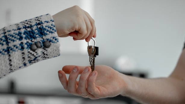Close-up picture of female hand in plaid jacket giving keys to new condominium to male hand with blurred kitchen appliances in background