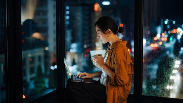Woman sitting by the window sill against illuminated city skyline at night, using laptop in the living room at home. Lifestyle and technology
