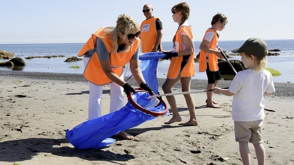 A little boy place some litter in a bag being held by a woman in an orange volunteer vest at the beach.