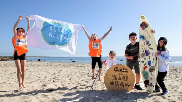 Children jumping and posing at a beach. Two on the left are holding a flag while three more are holding a surfboard.