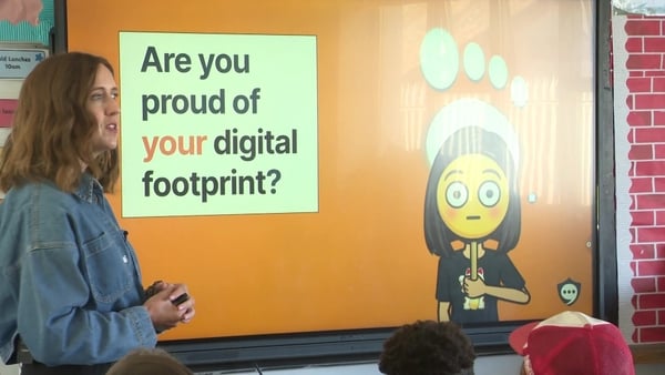 A woman stands in front of a screen giving a presentation at a school. The words Are you proud of your digital footprint appear there.