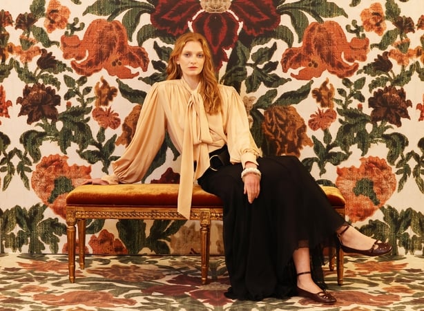 A woman poses in front of a floral backdrop wearing a loose blouse and long black skirt