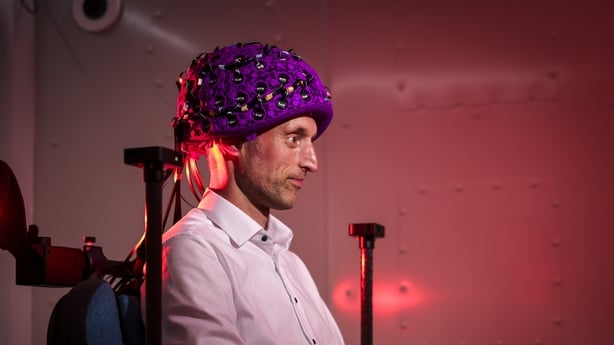 A man sits in a chair wearing the brain scanner on his head