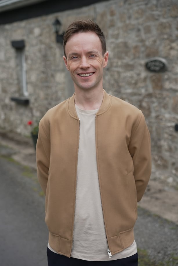 A man wearing a tan jacket stands in front of a stone house