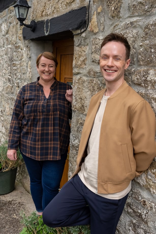 Two people stand in front of a stone house