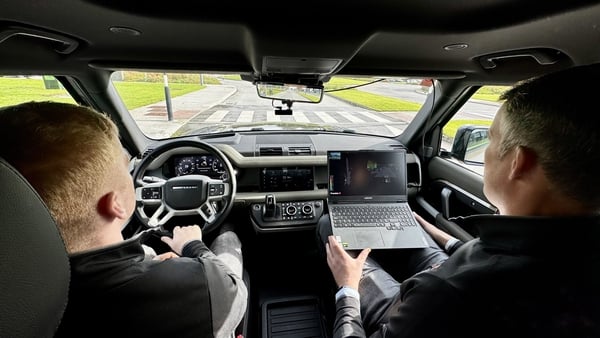 Two men sitting in a car. One man is driving and the other man is holding a laptop on his lap