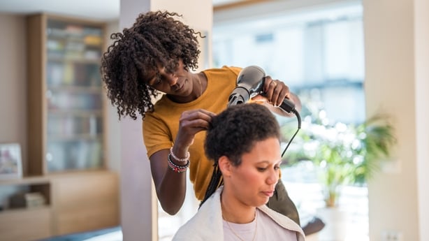 A mid adult Black female braids the hair of a mixed race female in a cozy home setting.