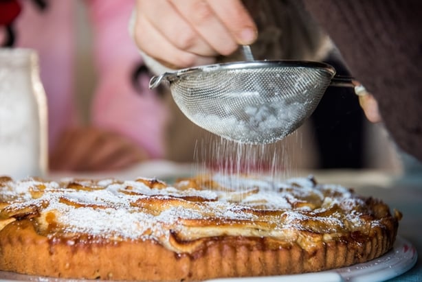 Close up of a child helped by her grandmother sifting powdered sugar using a sieve over a homemade apple tart