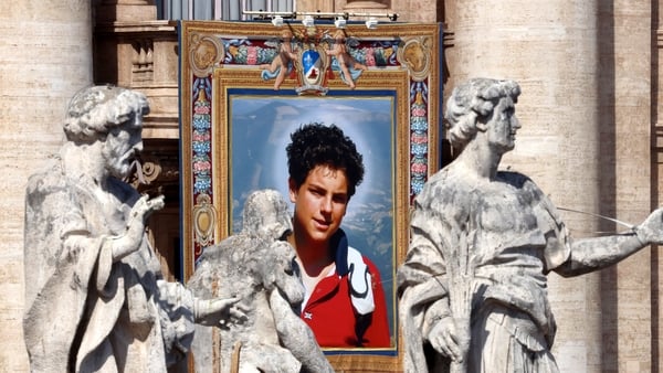A tapestry featuring Saint of the Catholic Church Carlo Acutis hangs from a balcony of St. Peter's Basilica in between three statues
