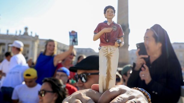 A model of Carlo Acutis held by hands while people watch in the background