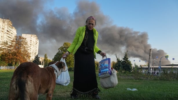 A woman speaks to a dog near the roadside as smoke rises in the background following a Russian drone and missile strike in Kyiv