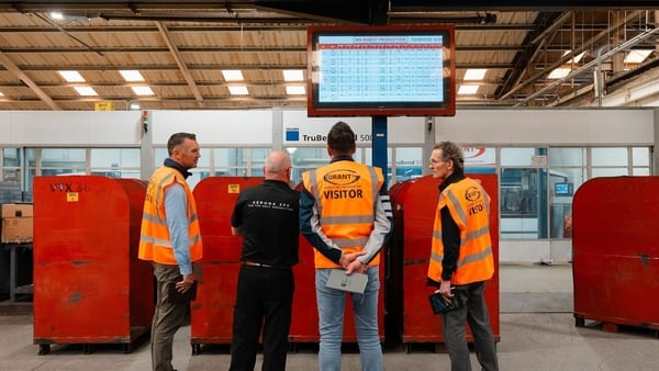 Four men, three of which are wearing orange high-vis jackets, look at a screen in a factory