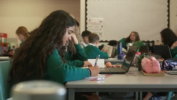A young girl takes notes wearing a green uniform sits in front of a laptop in a classroom. Other young students are in the background.