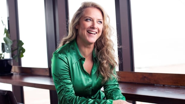 Soprano Sinéad Campbell Wallace sitting in front of a window, wearing a green silk blouse and smiling widely, looking off camera direction