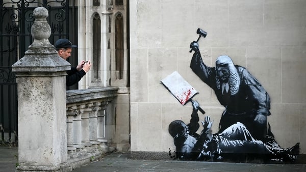 A person takes a photograph of an artwork by street artist Banksy, depicting a judge using a gavel to beat a protester using a placard as protection, painted on a exterior wall of the The Royal Courts of Justice, Britain's High Court, in London on Septemb