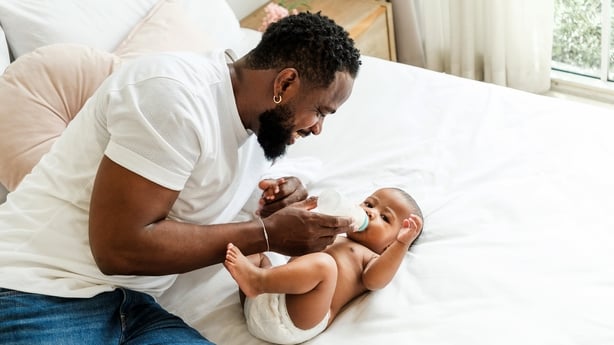 A father joyfully feeds his baby on a cozy bed, highlighting a moment of bonding and care. Happy diversity family and newborn bathing concept.