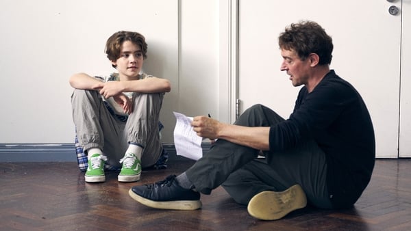 A man reads a script with a young boy on a TV set. Both are sitting on the floor.