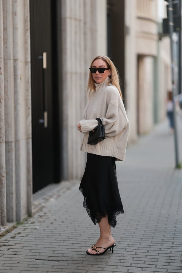 BERLIN, GERMANY - AUGUST 20: Sonia Lyson seen wearing Celine black cat-eye sunglasses, COS relaxed oversized beige turtleneck sweater in a chunky knit with dropped shoulders, Zara black asymmetrical slip dress featuring delicate lace trim along the hem, Chanel small black quilted top-handle Kelly ba