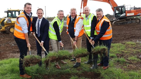 Group of men mostly in high vis vests digging in a field with construction vehicles in the background