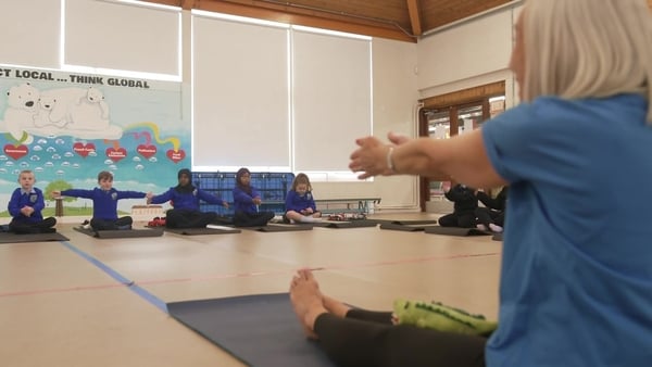 A woman, the foreground, shows schoolkids how to do yoga in the school hall.