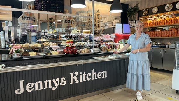 A woman stands in her café showing off the displayed cakes