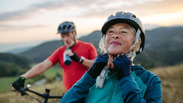 Portrait of beautiful senior couple on bike ride in autumn nature. Taking break, drinking water from sport bottle and fastening helmet.