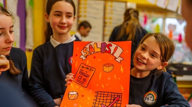A young girl in a school uniform shows off a poster she made to two other school girls with the word sales written on it and some charts drawn.