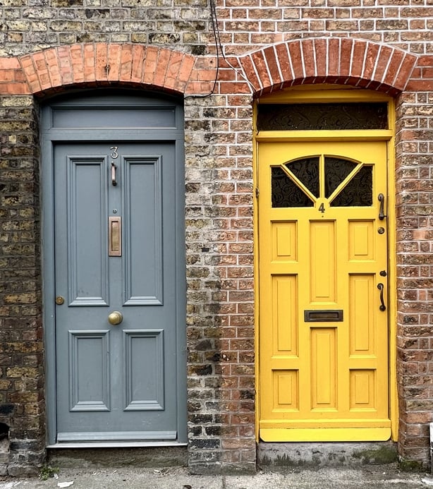 Two wood doors on brick wall with brick arch above. One grey door with panels, transom, centered door knocker, mail slot and door knob. One yellow door with panels and paned window and transom with door knocker and handle on right side. Gritty, urban feeling.