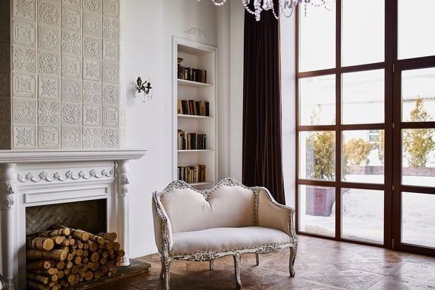 Luxurious living room featuring a white ornate fireplace, elegant upholstered sofa, built-in bookshelf, large wooden-framed windows, and classic tile accents, evoking warmth and sophistication.