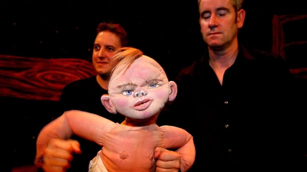A puppet in the foreground with two people in a dark background.