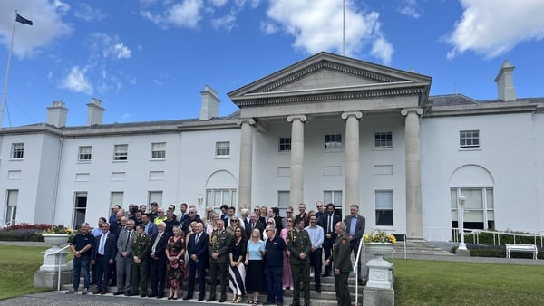 A group of people stand on the steps outside Áras an Uachtaráin on a sunny day.