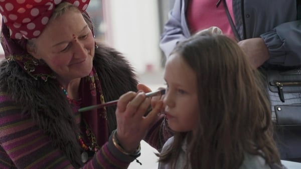 A woman with a red headband paints the face of a young girl sitting in a seat.
