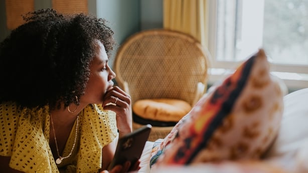 A woman looks as though she is reflecting on life as she gazes out of a bedroom window. She lies on her stomach on a bed, and touches her chin with her hand. She is holding a smart phone but isn't looking at it.